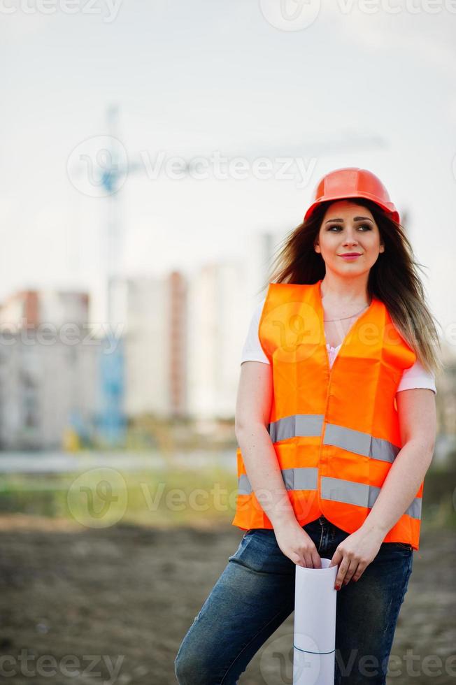 Engineer builder woman in uniform waistcoat and orange protective helmet hold business paper against new buildings with crane. photo