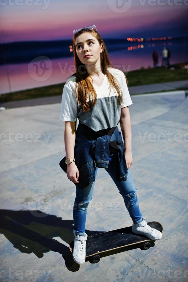 joven adolescente urbana con monopatín, gafas, gorra y jeans rotos en el parque de patinaje por la noche. foto