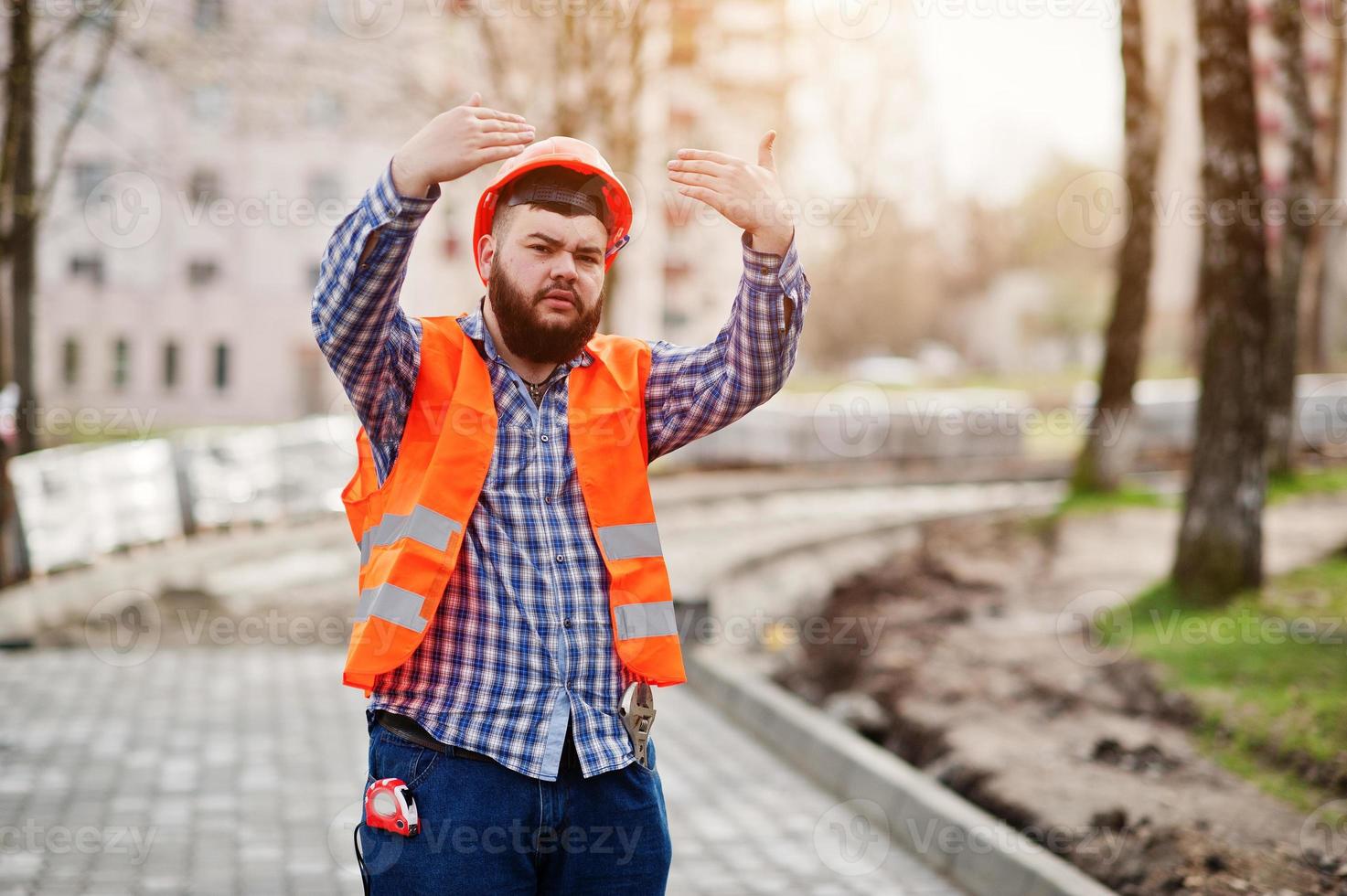 Portrait of brutal beard worker man suit construction worker in safety orange helmet against pavement with showing arms. photo