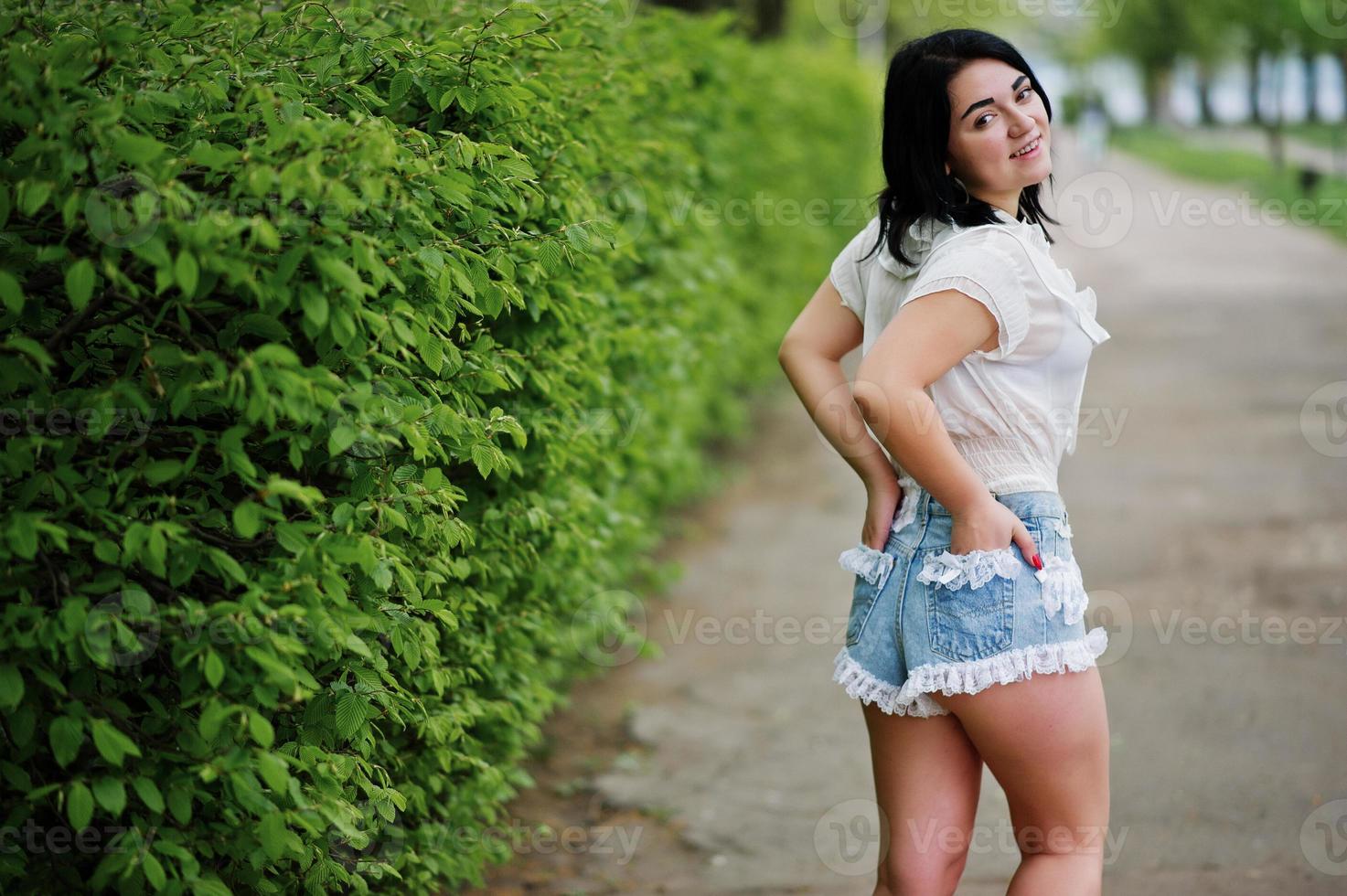 Back of sexy brunette girl on women's jeans shorts and white blouse against green spring bushes. photo
