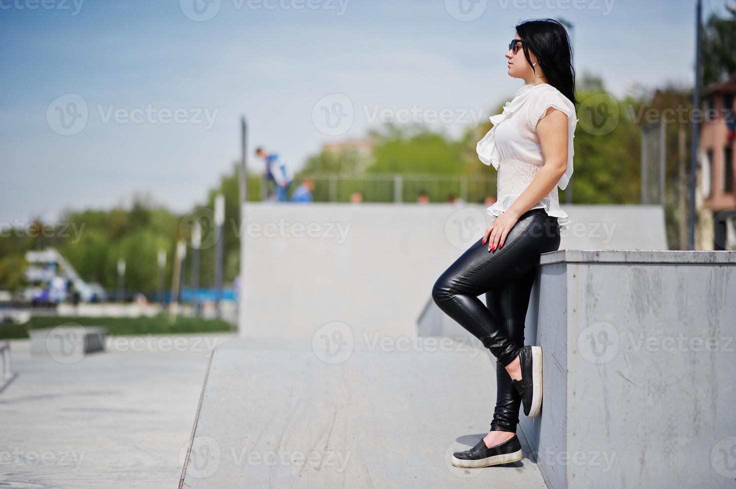 chica morena con pantalones de cuero de mujer y blusa blanca, gafas de sol, posada contra el parque de patinaje. foto