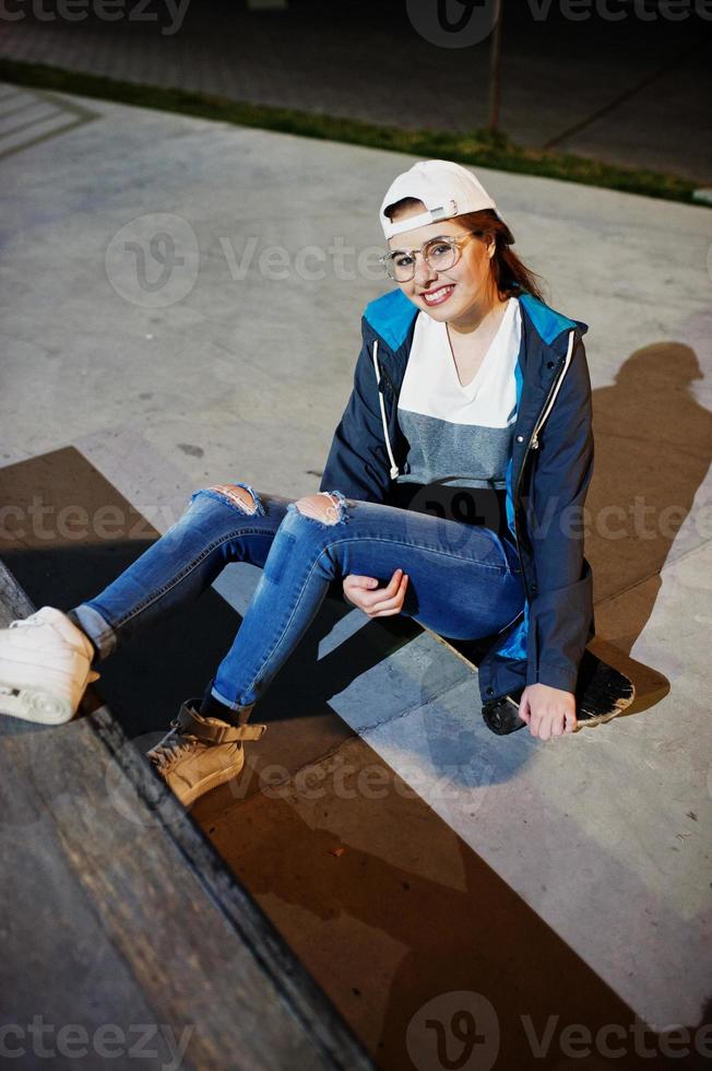 Young teenage urban girl with skateboard, wear on glasses, cap and ripped jeans at skate park on the evening. photo