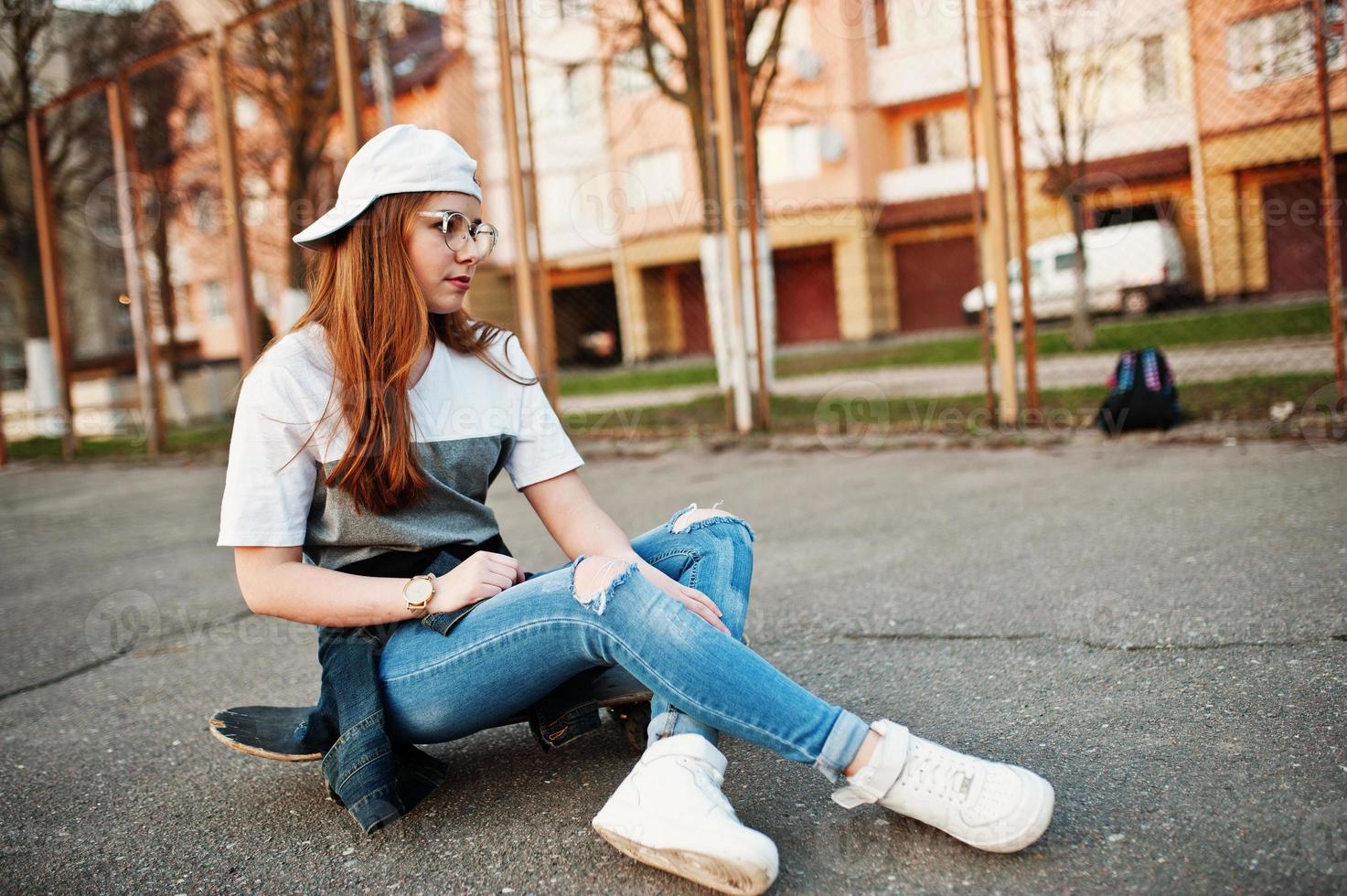 joven adolescente urbana con monopatín, gafas, gorra y jeans rotos en el campo de deportes del patio. foto