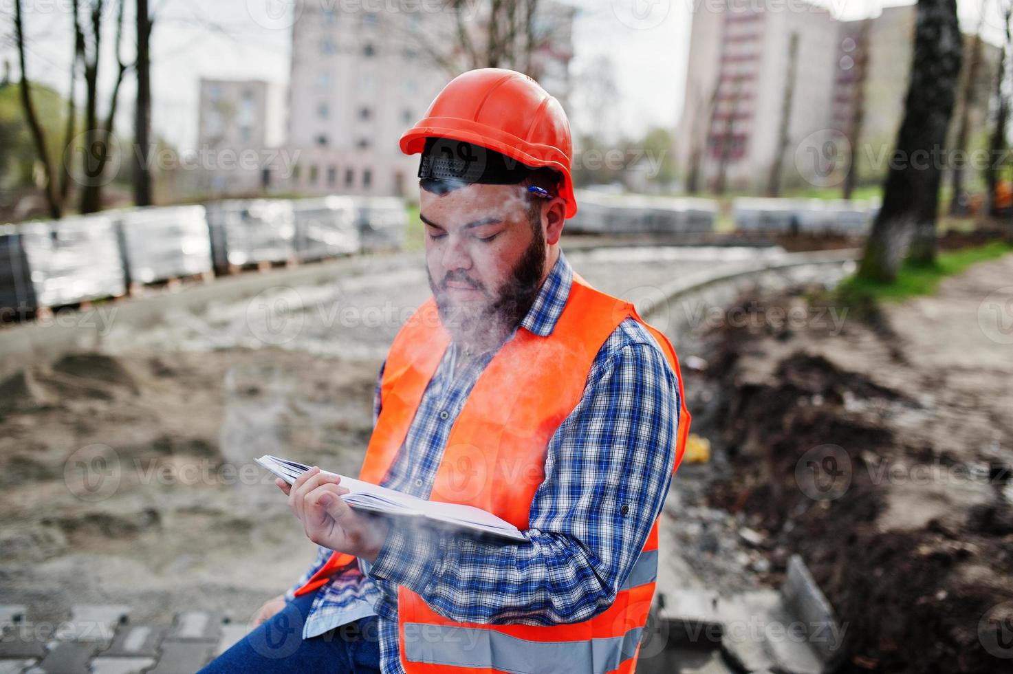 Brutal beard smoking worker man suit construction worker in safety orange helmet sitting on pavement, break at work, and read working notebook entries. photo