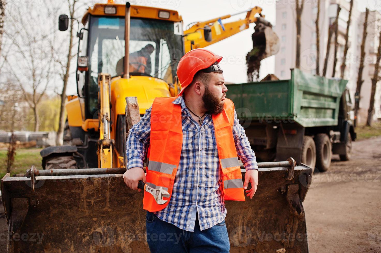 Brutal beard worker man suit construction worker in safety orange helmet, against traktor with adjustable wrench at hand. photo