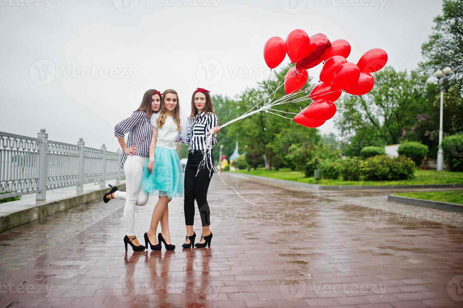 atractiva novia posando con sus tres encantadoras damas de honor con globos rojos en forma de corazón en el pavimento con un lago al fondo. despedida de soltera. foto