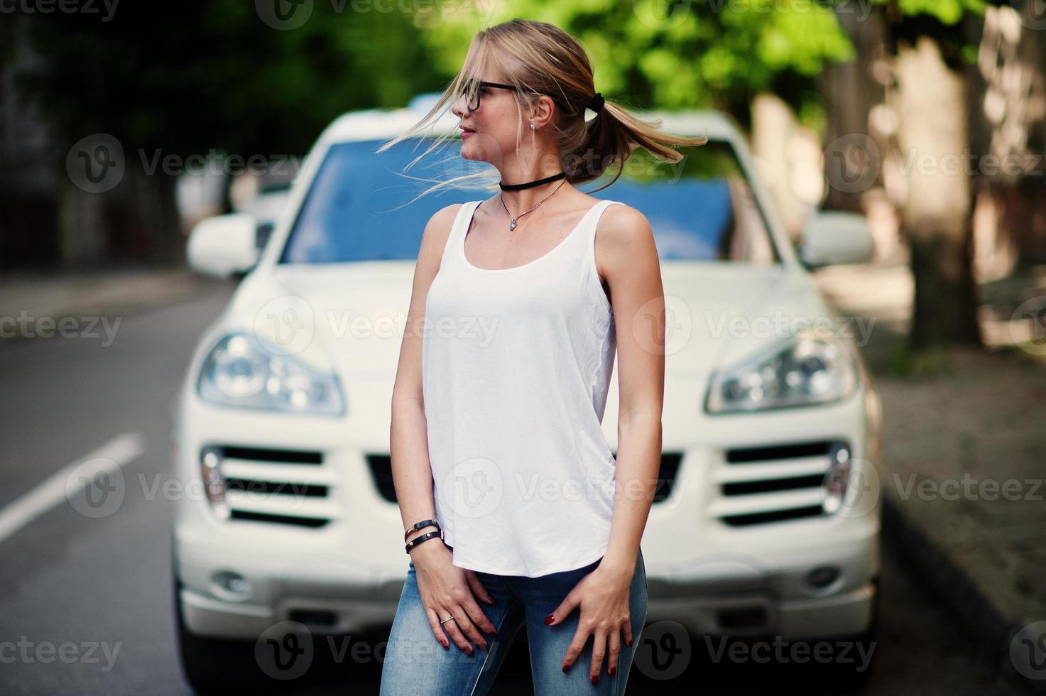 elegante mujer rubia vestida con jeans, gafas, gargantilla y camisa blanca contra un auto de lujo. retrato de modelo urbano de moda. foto