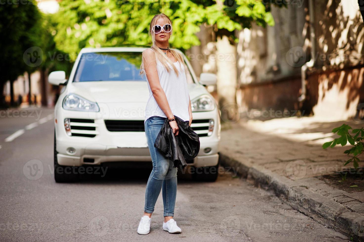 elegante mujer rubia vestida con jeans, gafas de sol, gargantilla y camisa blanca contra un auto de lujo. retrato de modelo urbano de moda. foto