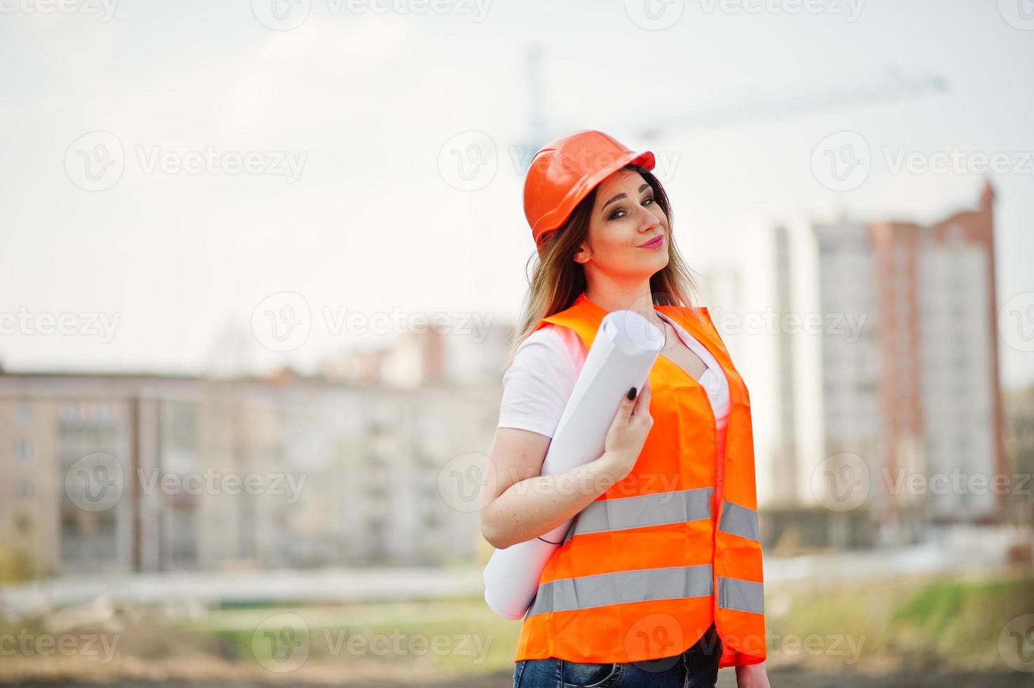 Engineer builder woman in uniform waistcoat and orange protective helmet hold business paper against new buildings with crane. photo