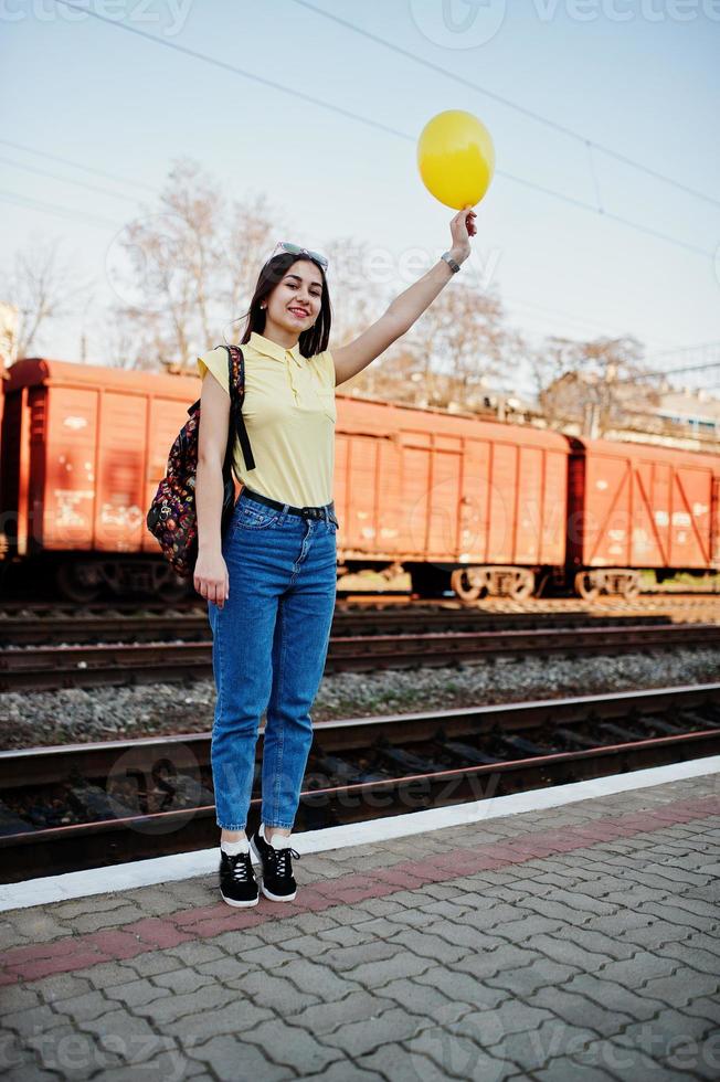Young teenage girl standing on the platform at the train station with ballon at hand, wear on yellow t-shirt, jeans and sunglasses, with backpack. photo