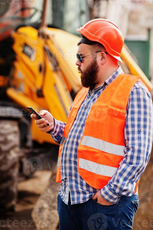 trabajador de barba traje de hombre trabajador de la construcción en casco naranja de seguridad, gafas de sol contra tractor con teléfono móvil a mano. foto