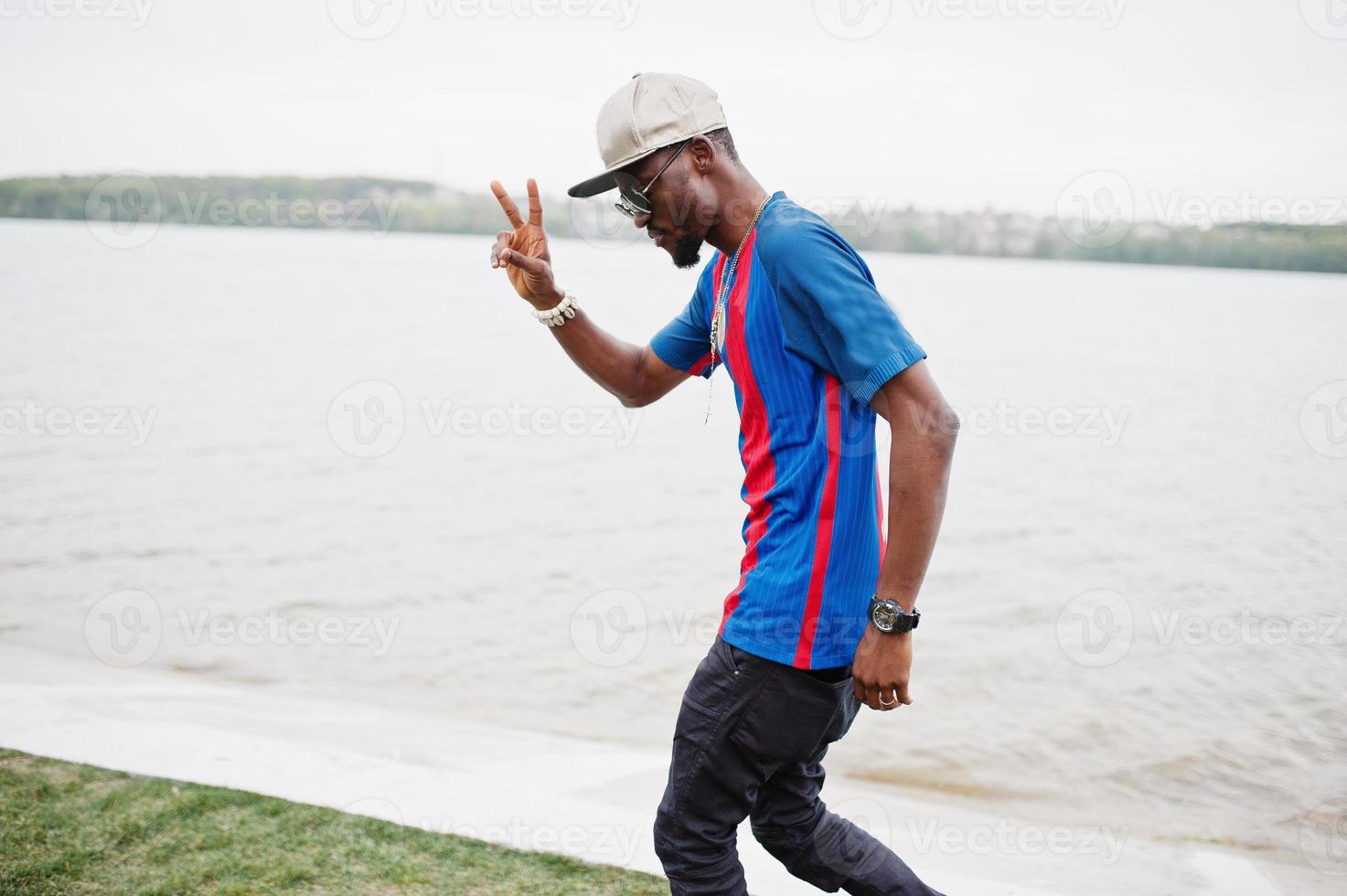 Stylish african american boy wear at cap, football t-shirt and sunglasses showing two fingers. Black sports man portrait. photo