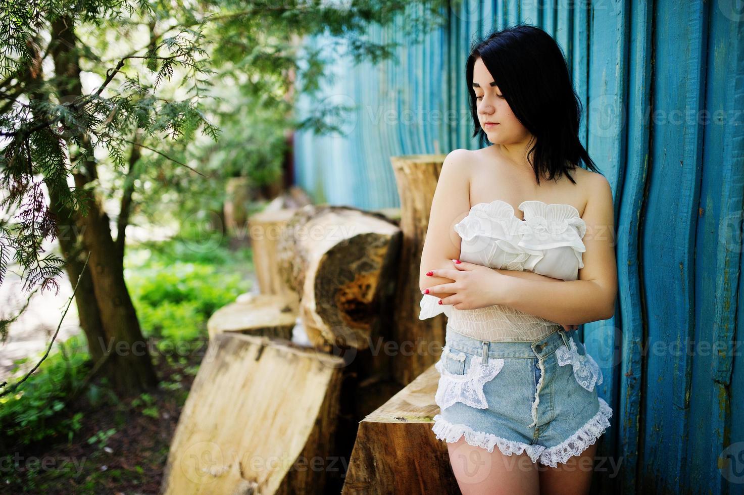 Portrait of sexy brunette girl on women's jeans shorts and white blouse against blue wooden house with stumps. photo