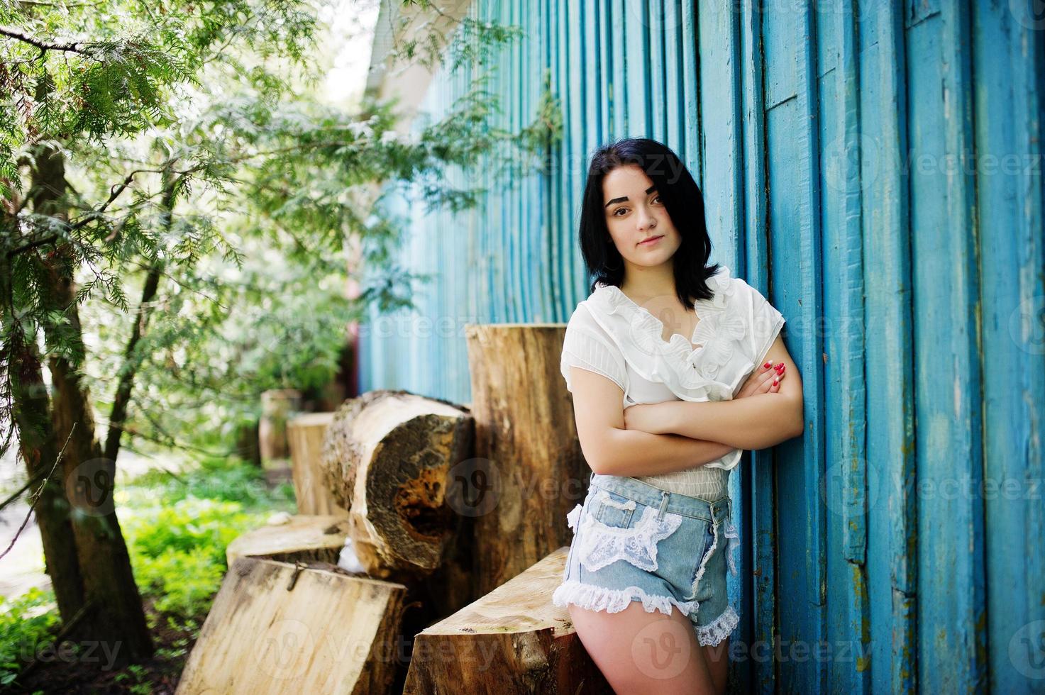 Portrait of sexy brunette girl on women's jeans shorts and white blouse against blue wooden house with stumps. photo