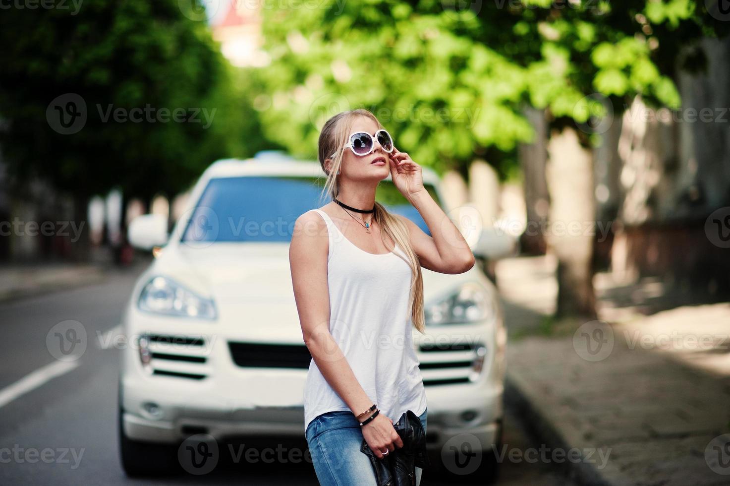 Stylish blonde woman wear at jeans, sunglasses, choker and white shirt against luxury car. Fashion urban model portrait. photo