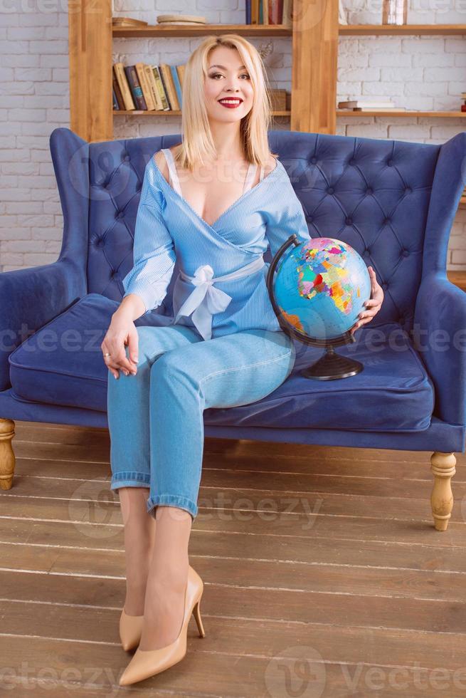 portrait of beautiful attractive cheerful blonde Slavic woman in blue jeans, blue blouse on the blue couch holding globe and dreaming about travelling photo