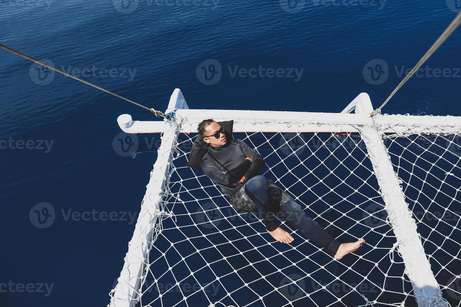 Asian man in wetsuit and sunglasses lying on catamaran net on vacation at Labuan Bajo photo