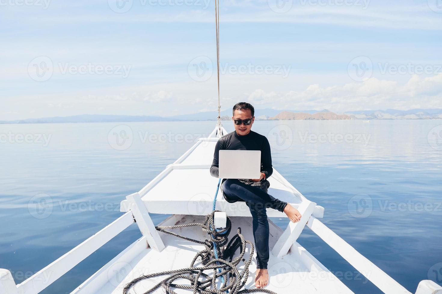 Asian man in wetsuit and sunglasses sitting on the boat and using a laptop on vacation at Labuan Bajo photo
