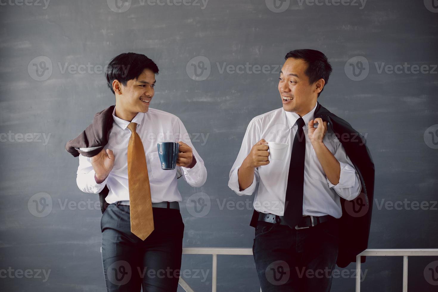 Two Asian businessmen in white shirts and ties chatting while holding coffee and coats over their shoulders photo