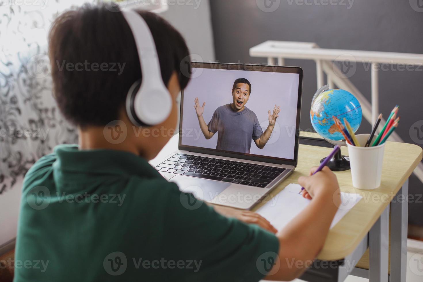 un colegial está escribiendo en un papel y mirando a su maestro en una computadora portátil foto