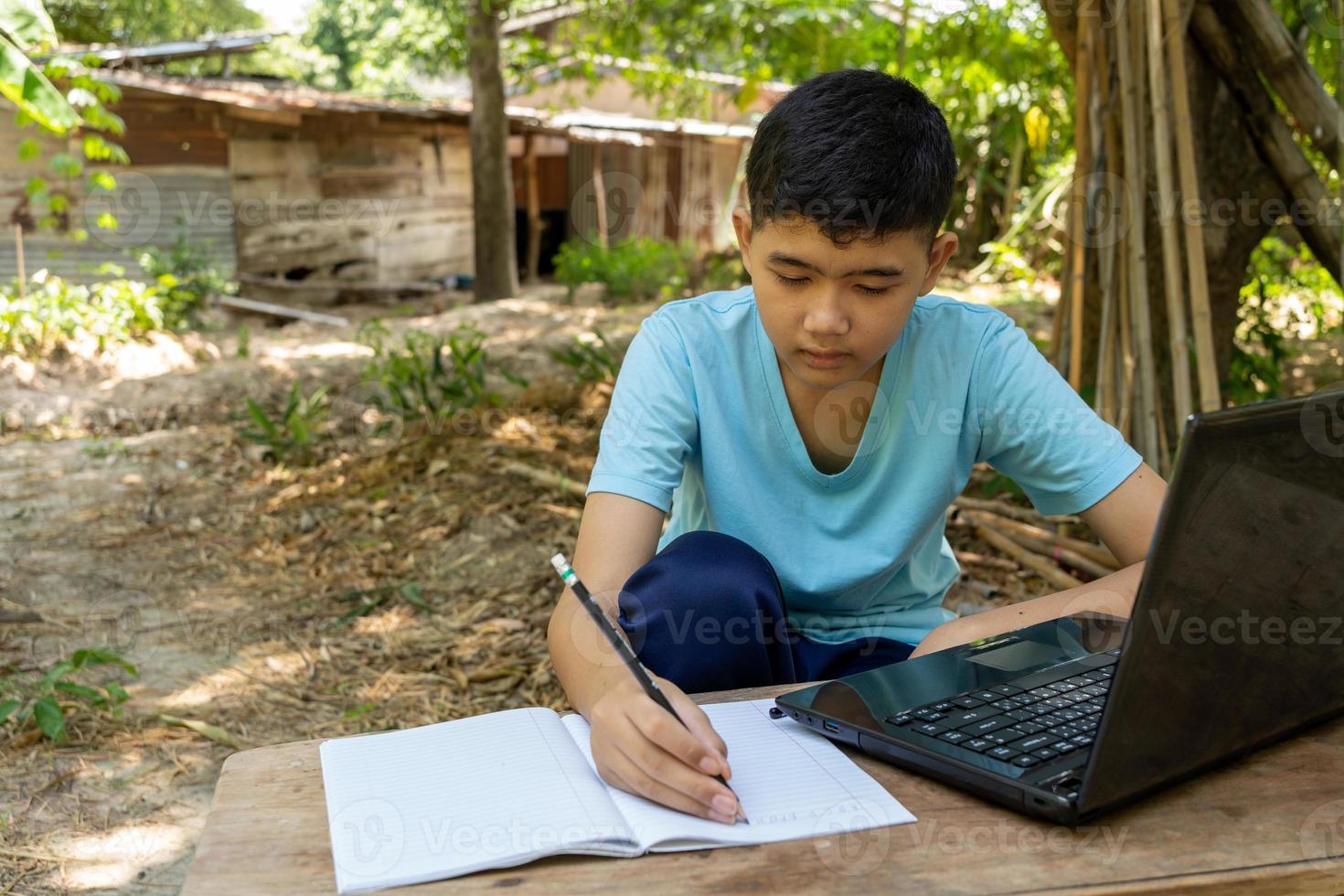 un niño está escribiendo en un cuaderno mientras estudia en línea con una computadora portátil en casa en el campo foto