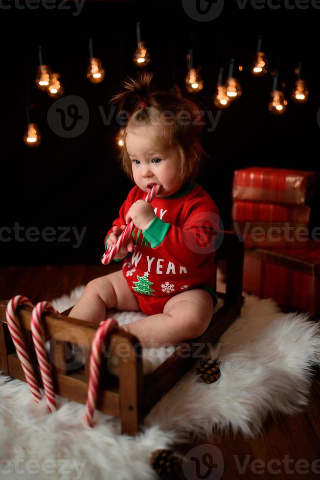 Una niña de 7 meses con un traje rojo de Navidad sobre un fondo de guirnaldas retro se sienta en una piel foto