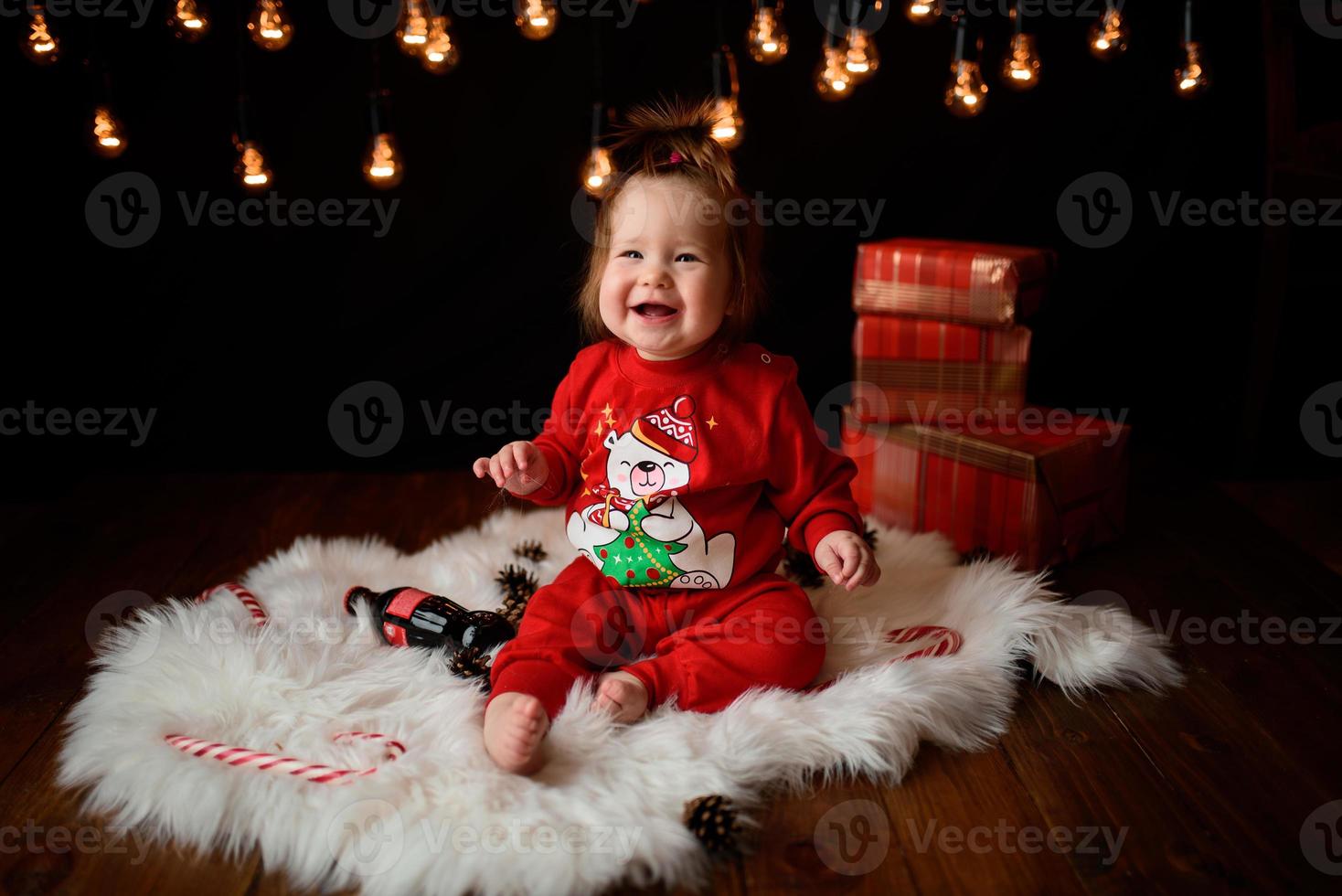 Una niña de 7 meses con un traje rojo de Navidad sobre un fondo de guirnaldas retro se sienta en una piel foto