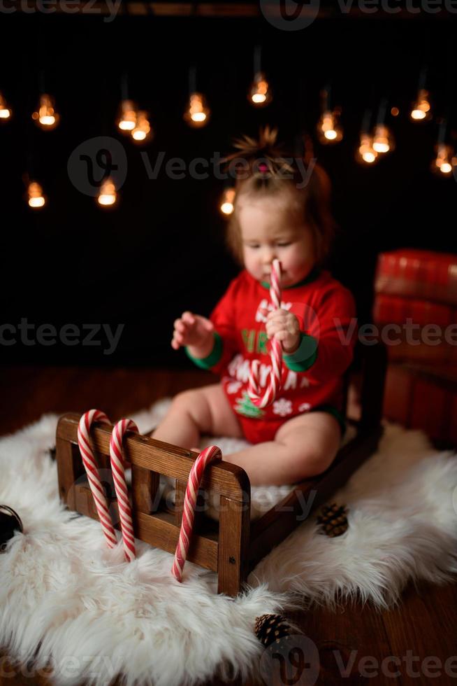 Una niña de 7 meses con un traje rojo de Navidad sobre un fondo de guirnaldas retro se sienta en una piel foto