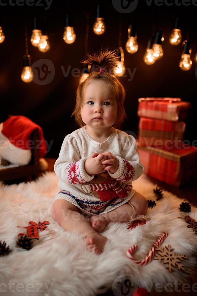 Una niña de 7 meses con un traje rojo de Navidad sobre un fondo de guirnaldas retro se sienta en una piel foto