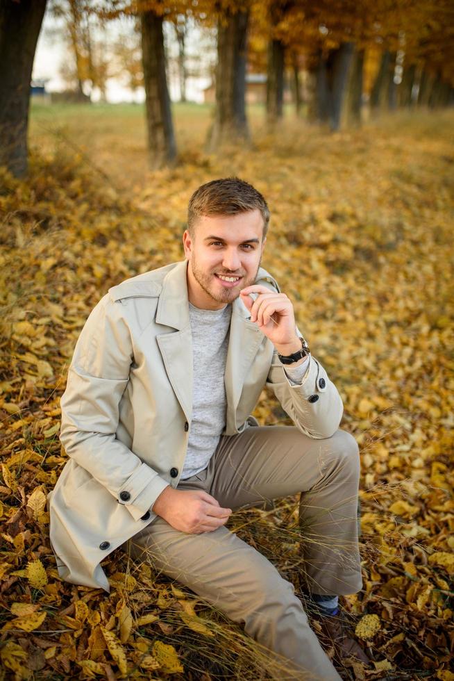 Portrait of a young man in a coat on a background of autumn trees. A man sits on the ground. photo