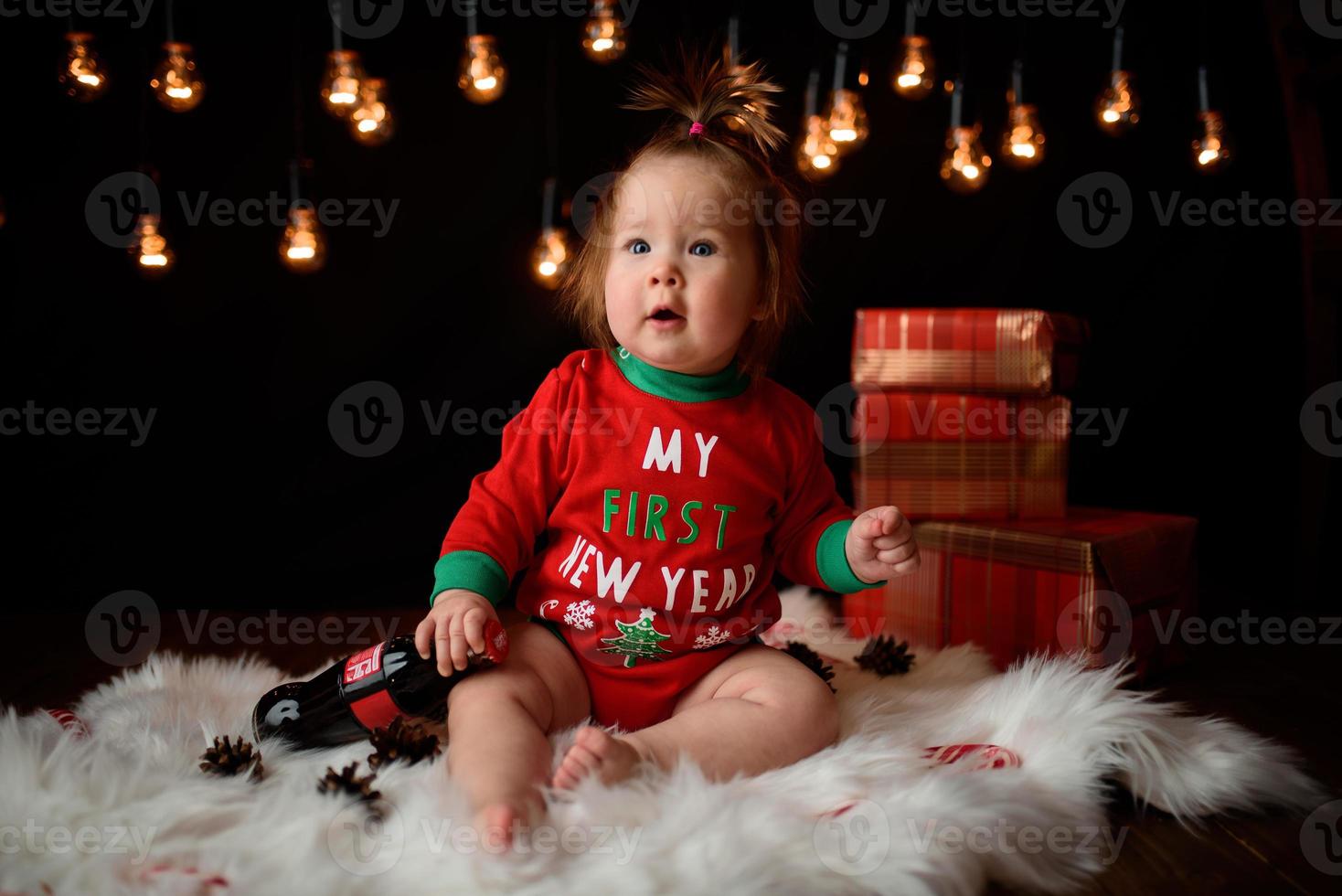 Una niña de 7 meses con un traje rojo de Navidad sobre un fondo de guirnaldas retro se sienta en una piel foto