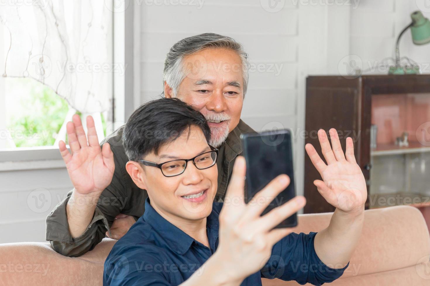 Happy senior asian father and adult son using smart phone talking on video call in living room photo