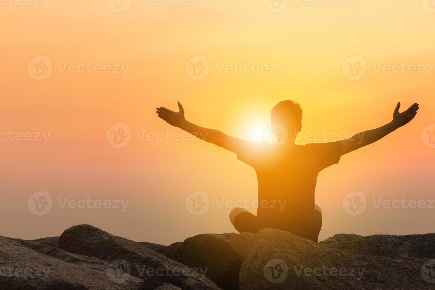 silueta de hombre sentado con las manos abiertas sobre una piedra mirando el sol dorado. puesta de sol en el cielo nocturno en el fondo de la playa, deporte y disfrutar de la vida en el concepto de mar. foto