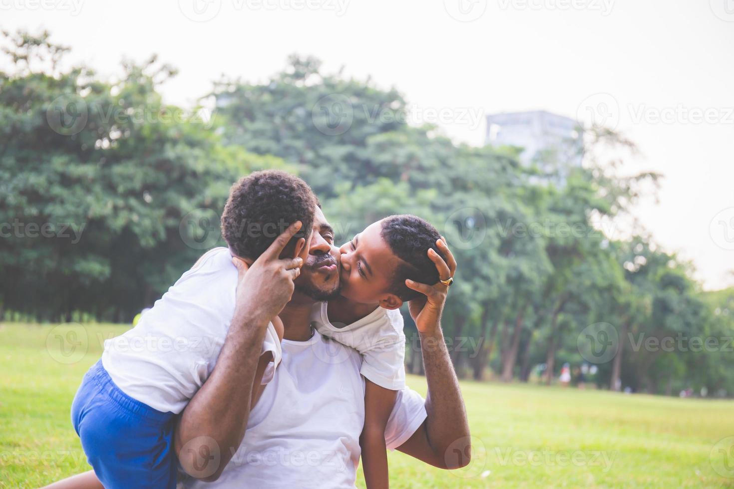 niño lindo africano en casual besando a papá en el parque. feliz padre afroamericano jugando al aire libre con su hijo, alegre concepto de familia negra. foto