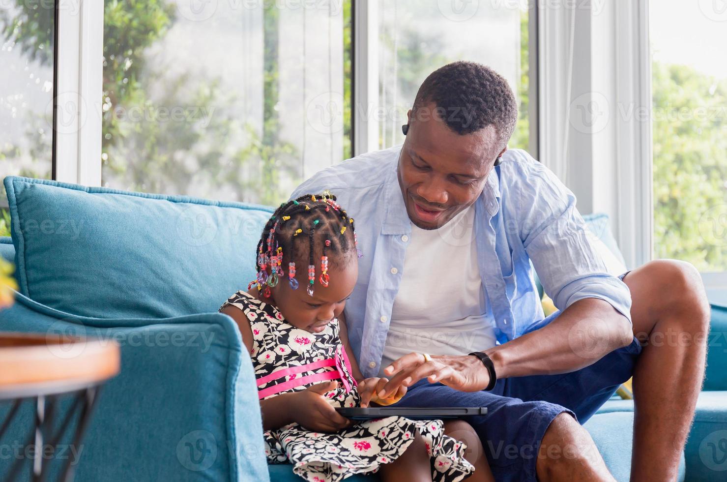 alegre padre afroamericano e hija jugando en la sala de estar, linda niñita y papá sentados en el sofá y jugando en la tableta, conceptos familiares de felicidad foto