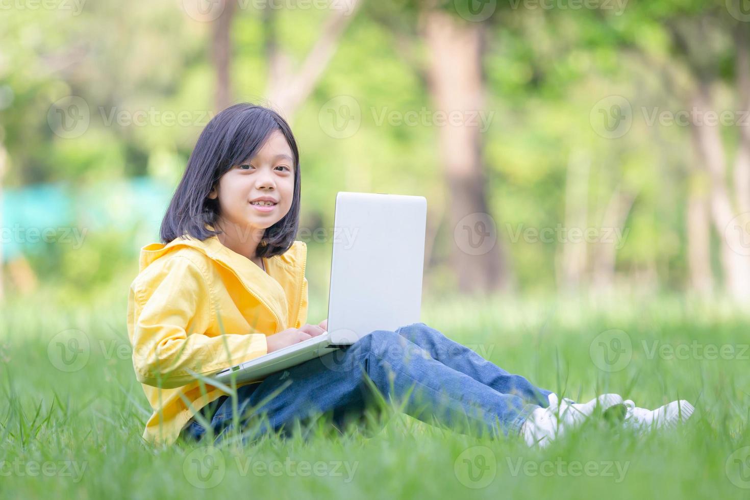 Asian  kid girl sitting on the grass studying with laptop in the park, Outdoor learning concepts photo