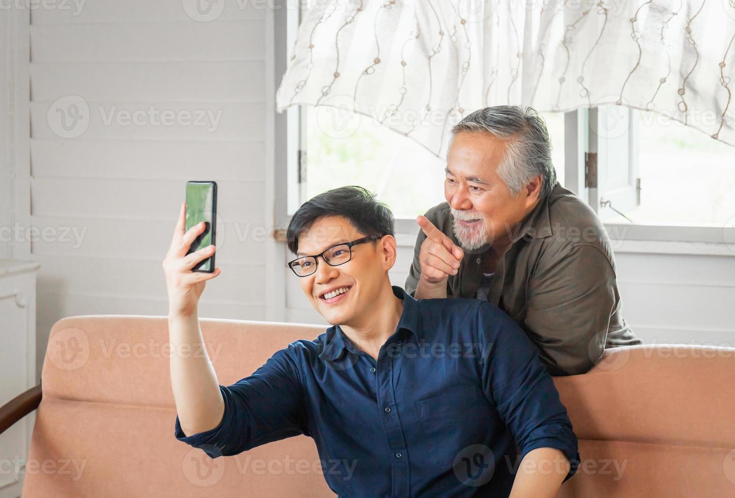 Cheerful senior asian father and adult son using smart phone talking on video call in living room photo