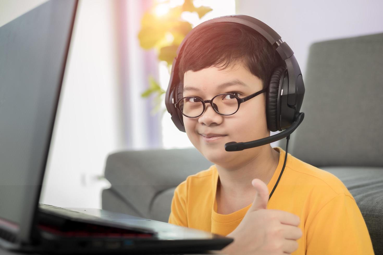 joven y lindo niño asiático de 10 años con auriculares sentado en la sala de estar y usando una computadora portátil para estudiar a distancia en línea y pulgar arriba con cara feliz durante el brote de coronavirus foto