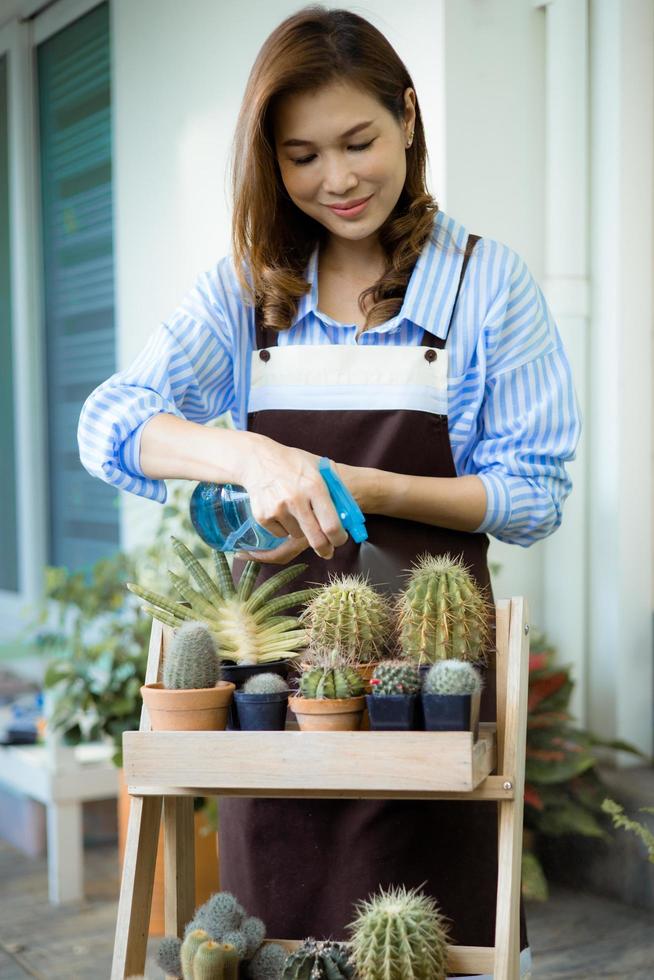 linda ama de casa asiática con delantal sosteniendo un rociador de agua y regando la maceta de pequeñas plantas y cactus con cara sonriente y feliz con el hobby en casa en el tiempo libre foto