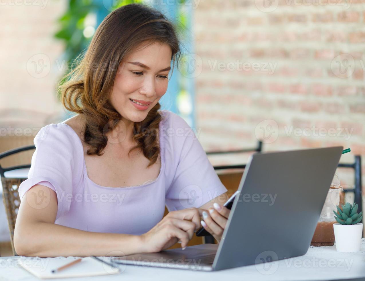 The beautiful middle-aged woman sitting in food shop and working with laptop note computer and writing on memo paper. Freelancer job and work from anywhere concept photo