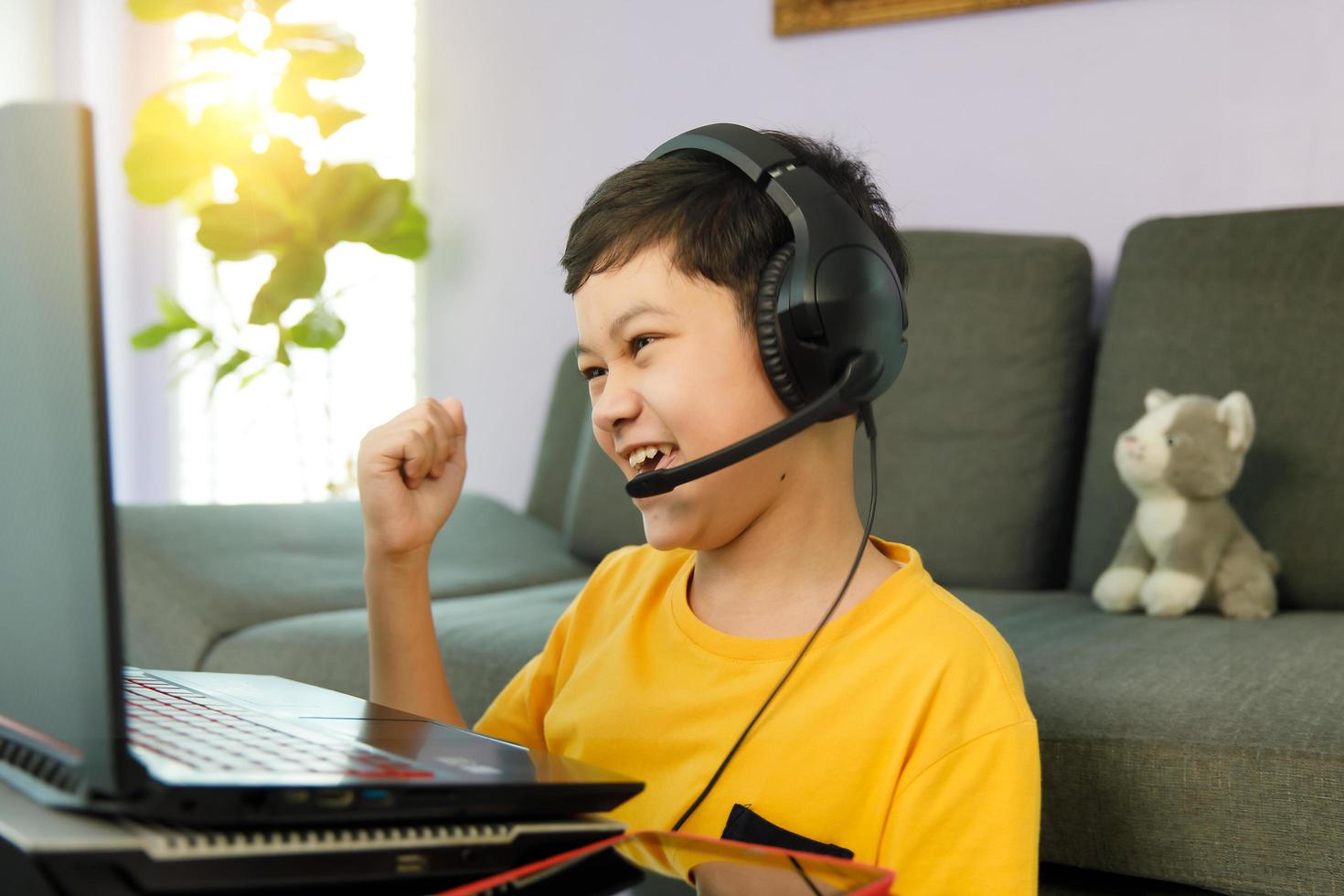 Joven y lindo niño asiático de 10 años con audífonos sentado en la sala de estar de su casa usando una computadora portátil para estudiar a distancia en línea y levantar el puño con entusiasmo y confianza en sí mismo foto