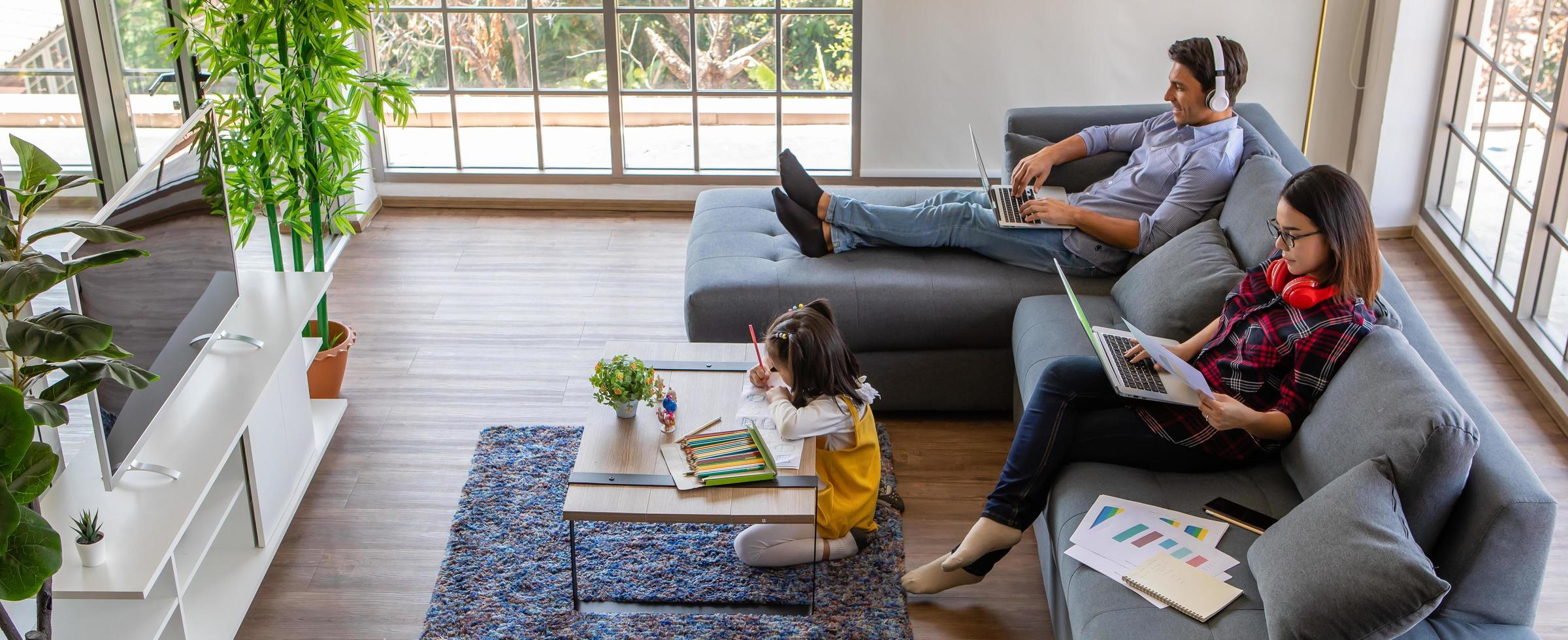 Multiracial family, father, mother, and little daughter stay together at home. Mom and dad working with laptop notebook computer while young girl does homework. Work from home and new normal concept photo