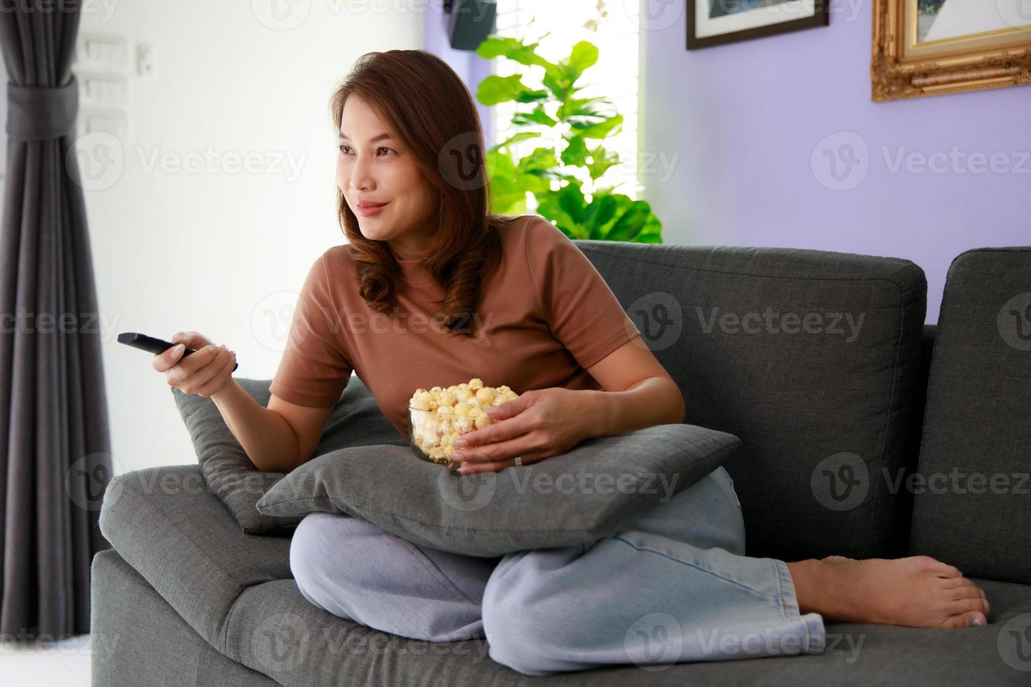 Mid adult Asian woman sitting on sofa in home living room holding bolw of popcorn and using remote control aiming to tv while watching in relax time photo