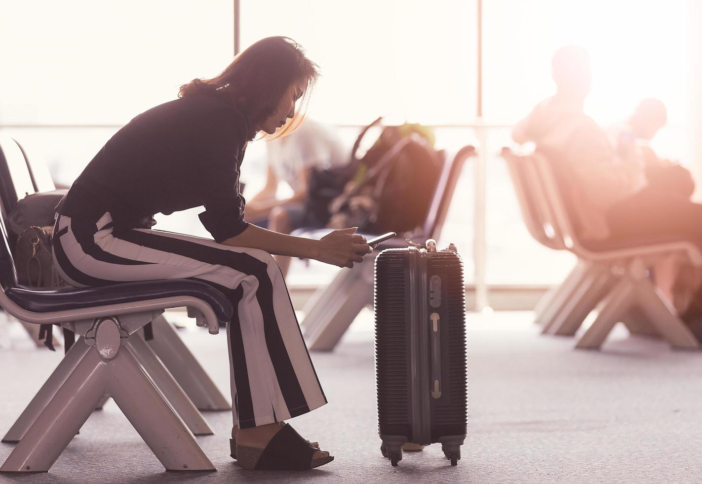 mujer sentada en el vestíbulo del aeropuerto usando un teléfono inteligente y mirando la pantalla mientras espera el tránsito. luz del sol brillando desde afuera foto