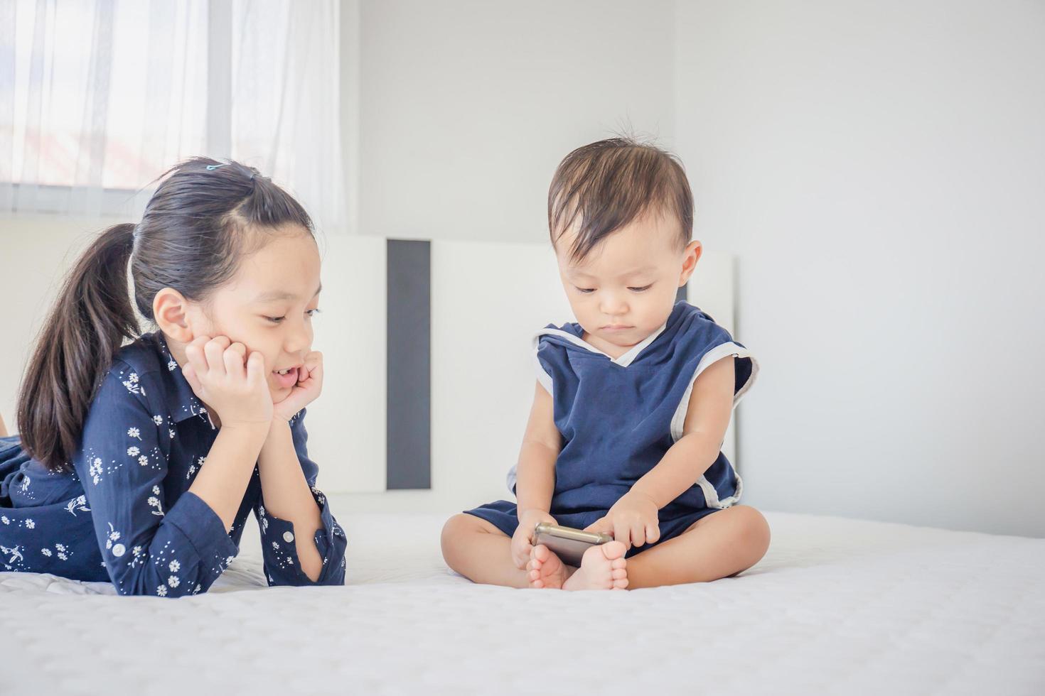 Sister and baby brother using smartphone, watching cartoons together on the bed, happy family and kids playing concept photo