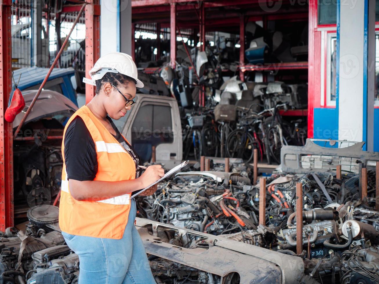 African woman warehouse automotive parts worker wears a safety helmet takes note on the clipboard among the old engines automotive spare parts. photo