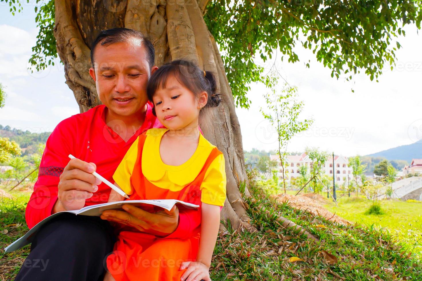 The father teaching daughter outside on filed under the tree. Father and daughter reading book washing hand under the tree. photo