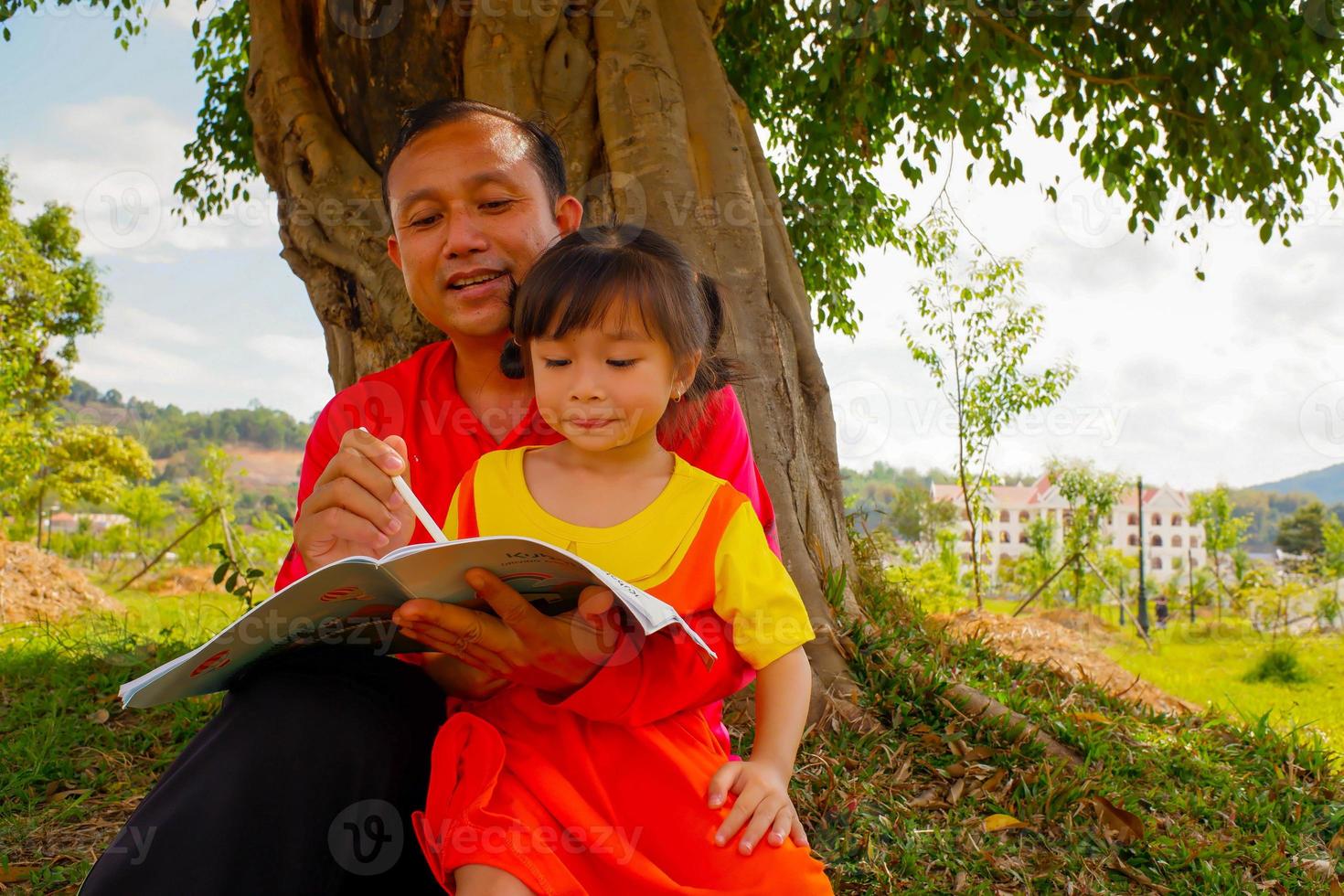 The father teaching daughter outside on filed under the tree. Father and daughter reading book washing hand under the tree. photo