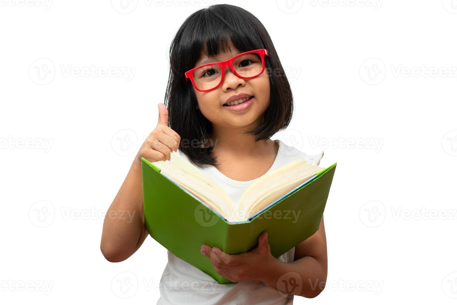 feliz niña preescolar asiática con gafas rojas sosteniendo un libro verde y pulgares hacia arriba sobre fondo blanco aislado. concepto de niño escolar y educación en primaria y preescolar, escuela en casa foto