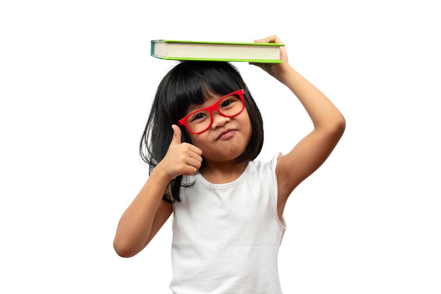 Happy Asian little preschool girl wearing red glasses holding a green book and thumbs up on white isolated background. Concept of school kid and education in elementary and preschool, home school photo